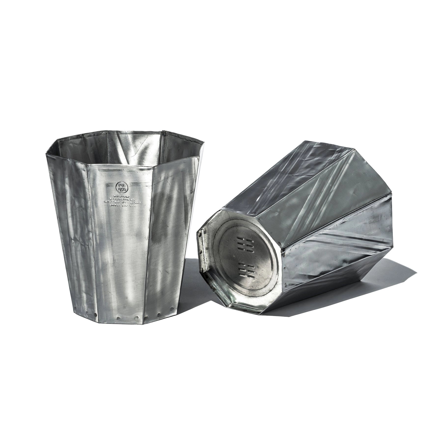 Two metallic Steel Trashcans from Faire, made of recycled steel with riveted seams and shiny silver surfaces—one upright, one on its side—are displayed against a white background, highlighting their industrial design.