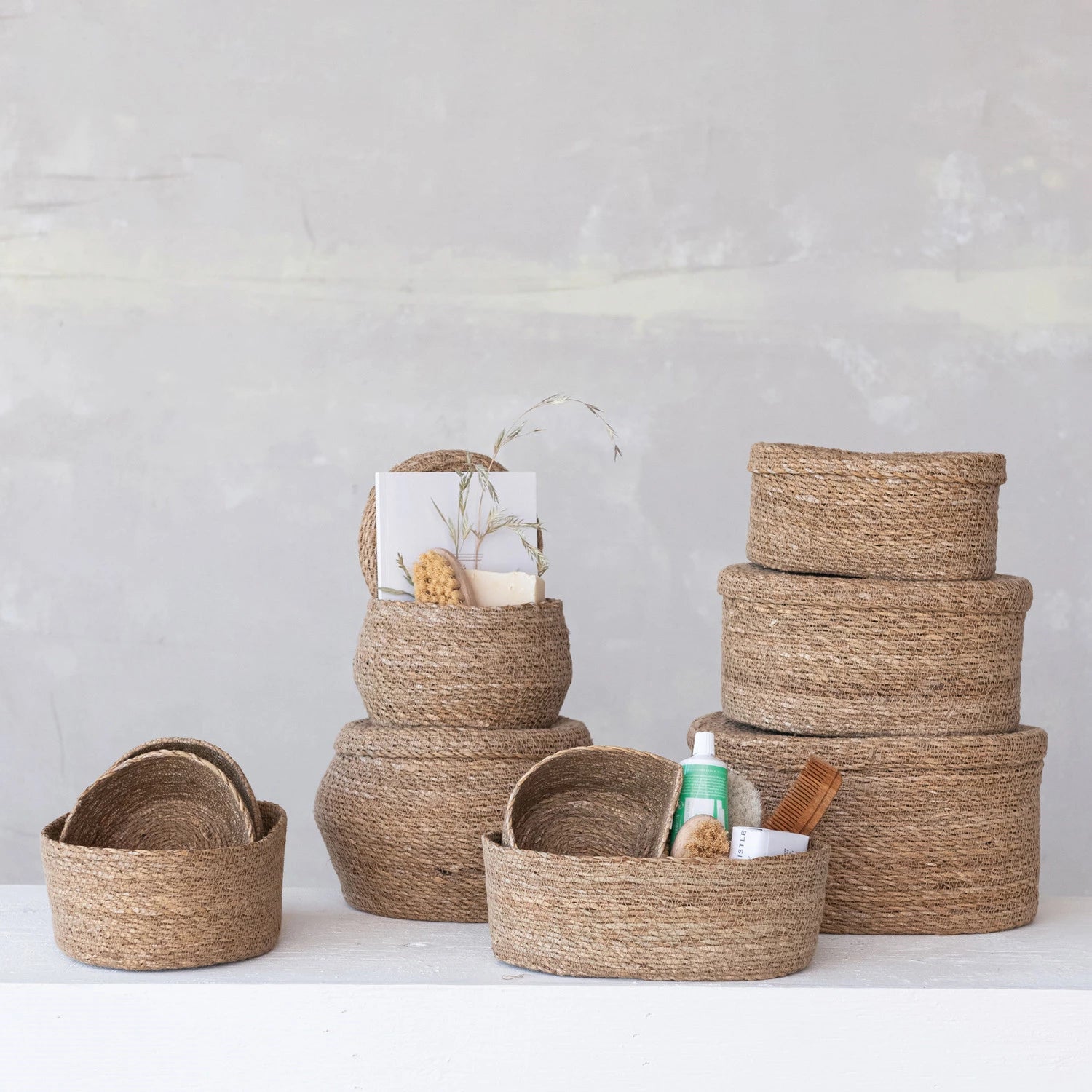 A set of Creative Co-op Sea Grass Basket (Long Oval) in different sizes, some stacked and others holding toiletries and brushes, displayed on a white surface with a light gray wall backdrop.