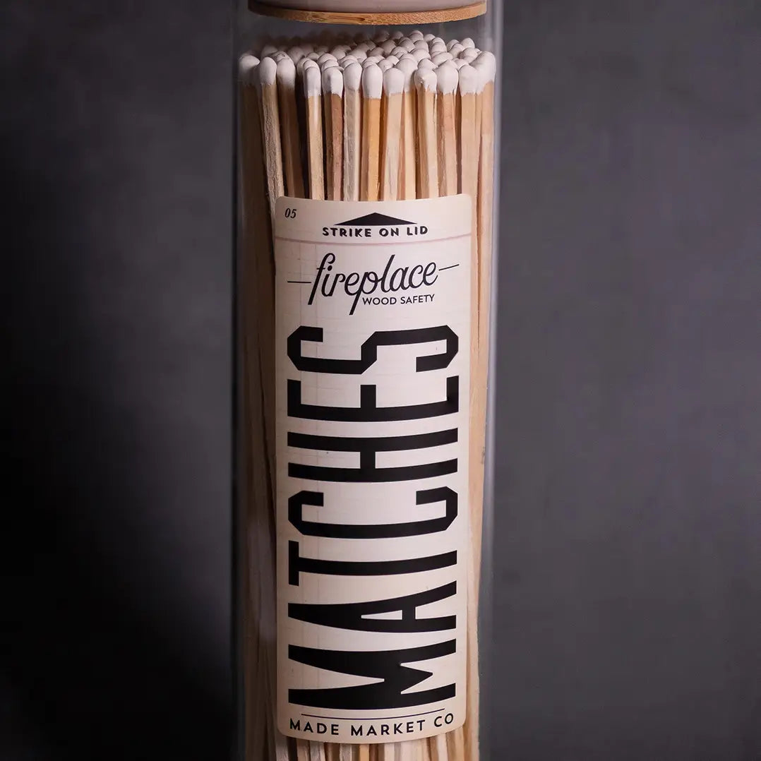 A close-up of a tall glass jar filled with long Fireplaces Apothecary Matches from Faire. The bold label details are visible, and the background is dark and plain.