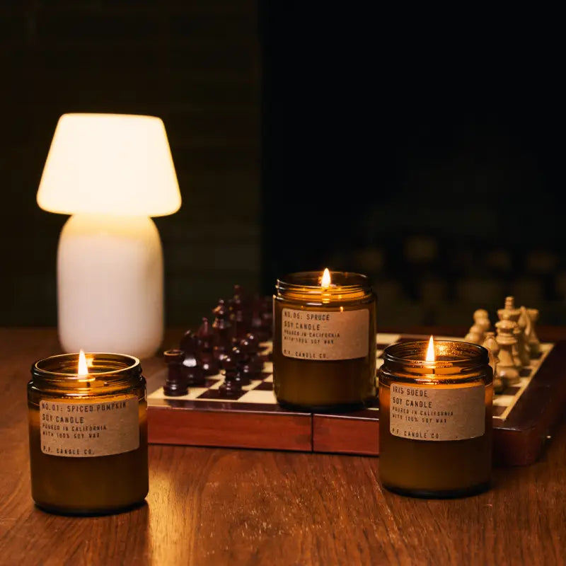 Three Faire PFC Candles, hand-poured soy wax in glass jars, rest on a wooden table beside a chessboard and glowing lamp, infusing the cozy space with inviting fine fragrance oils.