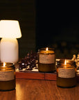 Three Faire PFC Candles, hand-poured soy wax in glass jars, rest on a wooden table beside a chessboard and glowing lamp, infusing the cozy space with inviting fine fragrance oils.