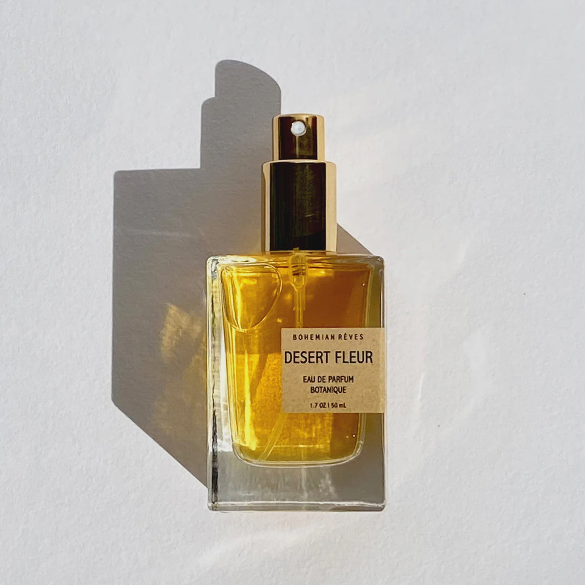 A square glass bottle of Faire's Desert Fleur Botanical Parfum Mist, filled with golden liquid and infused with Southwest Spirit, is set against a white background. The bottle casts a shadow, and the label describes hints of patchouli.