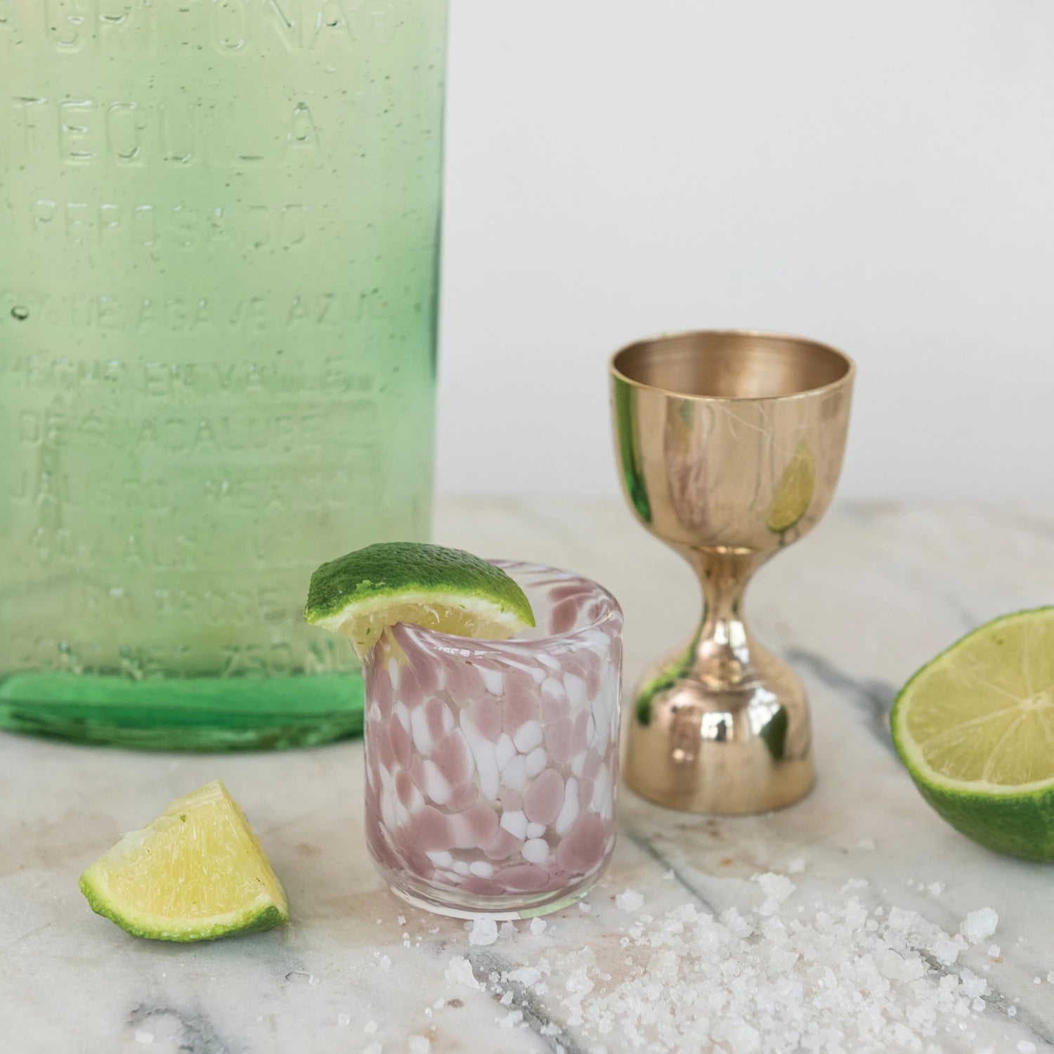 A marbled shot glass with a drink and lime wedge rests on marble next to the Creative Co-op Brass Double Sided Cocktail Jigger, a green bottle, lime slices, and coarse salt.