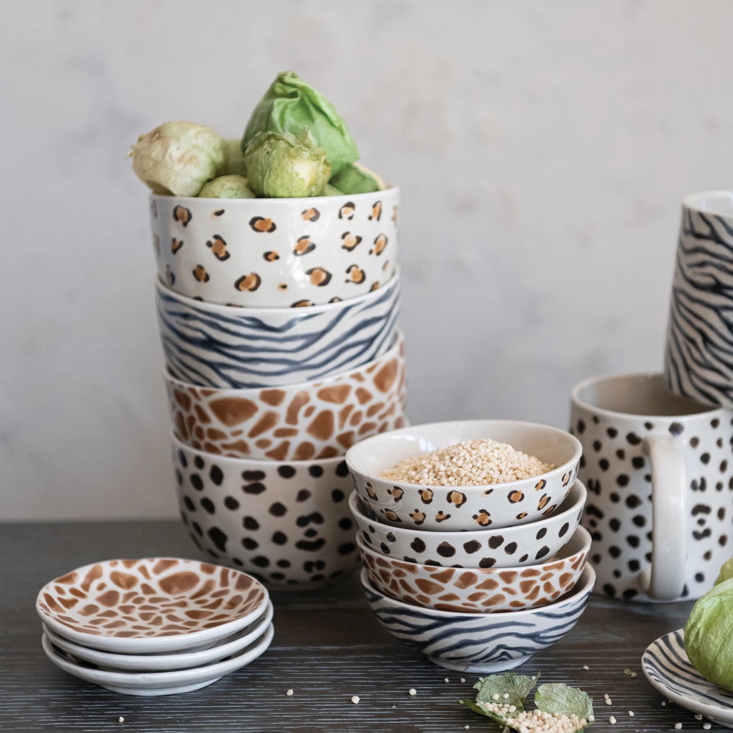 A Creative Co-op Bowl with Animal Print features leopard, giraffe, and zebra patterns. Shown with matching plates and a mug, plus serving dishes holding Brussels sprouts and grains on a dark surface scattered with food.