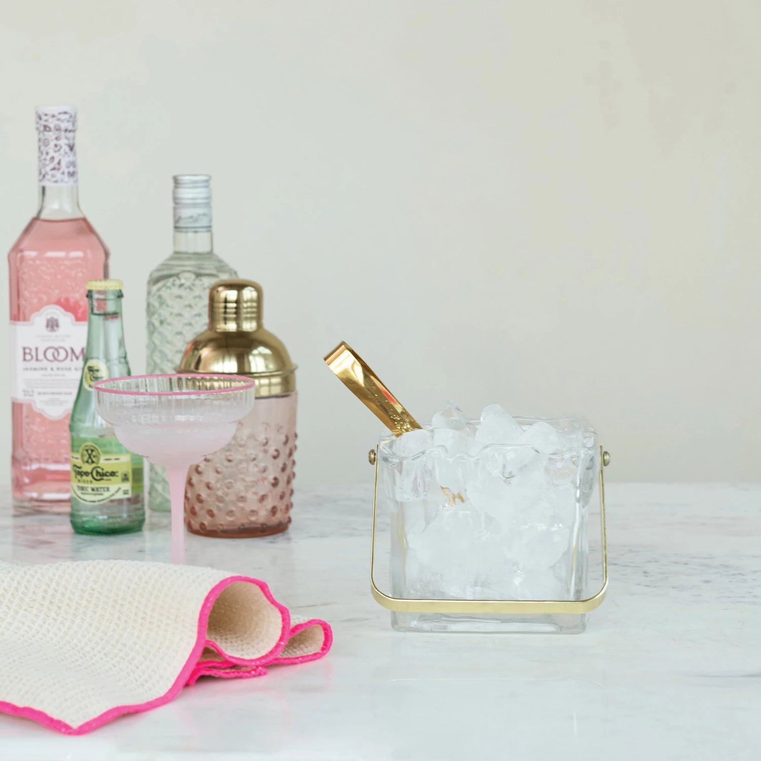 A marble countertop displays spirits, a cocktail glass, a gold shaker, a textured towel, and the Creative Co-op Glass Ice Bucket with Tongs featuring a textured design and a gold-finished handle.