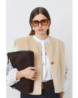 A woman with straight brown hair wears SUNCOO's Diego Vest over a white blouse, paired with blue jeans, gold earrings, and large square sunglasses. She holds a large brown clutch against a plain white background.
