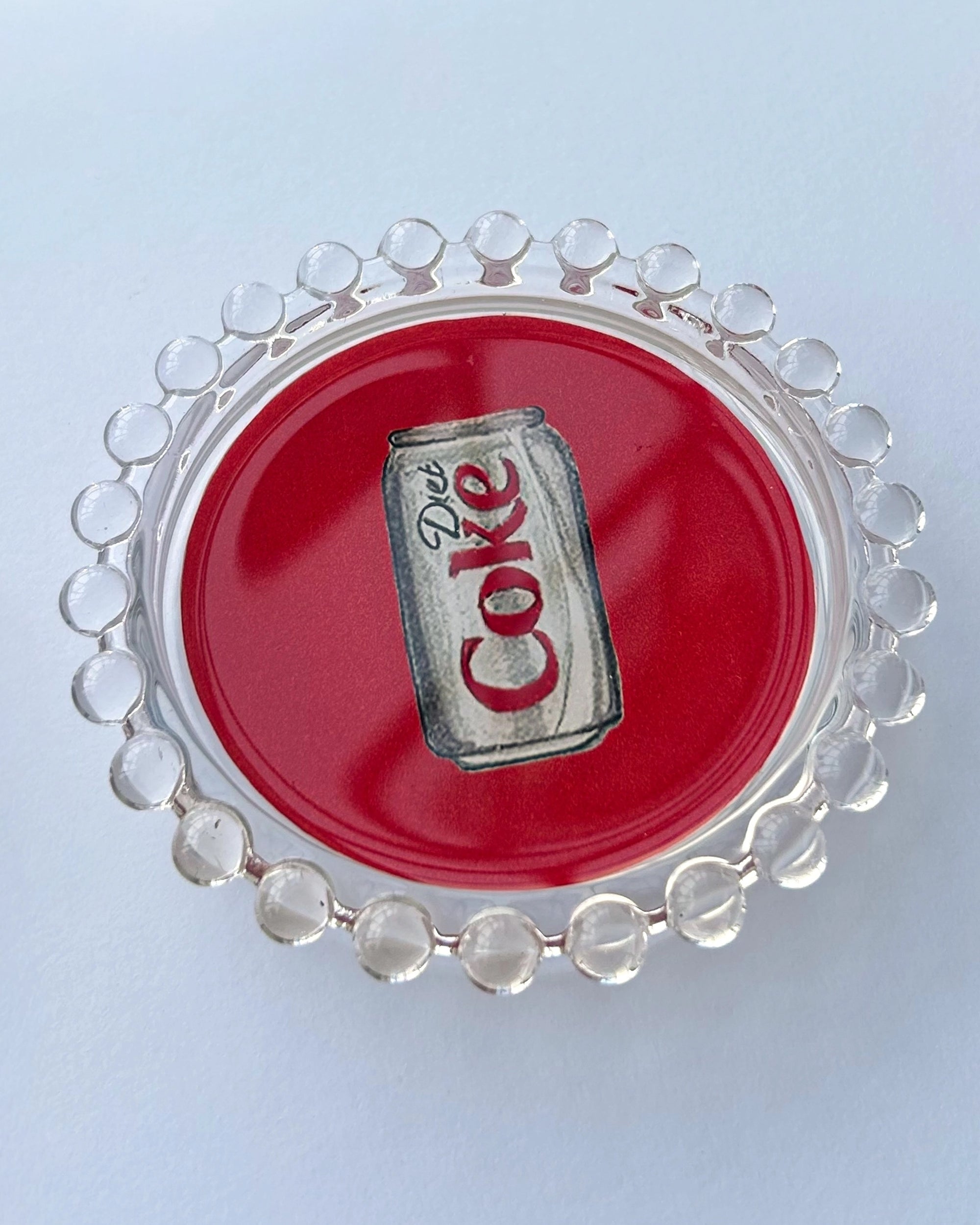 The Faire Diet Coke Dish is a clear glass plate with a beaded edge and a Diet Coke can illustration on a red background—ideal as a unique jewelry tray or playful housewarming gift.