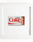 A framed artwork showcasing a miniature Diet Coke can label partially wrapped over a small matchbox with red-tipped matches, all against a pure white background in Match South style using the Art Square White Frame.