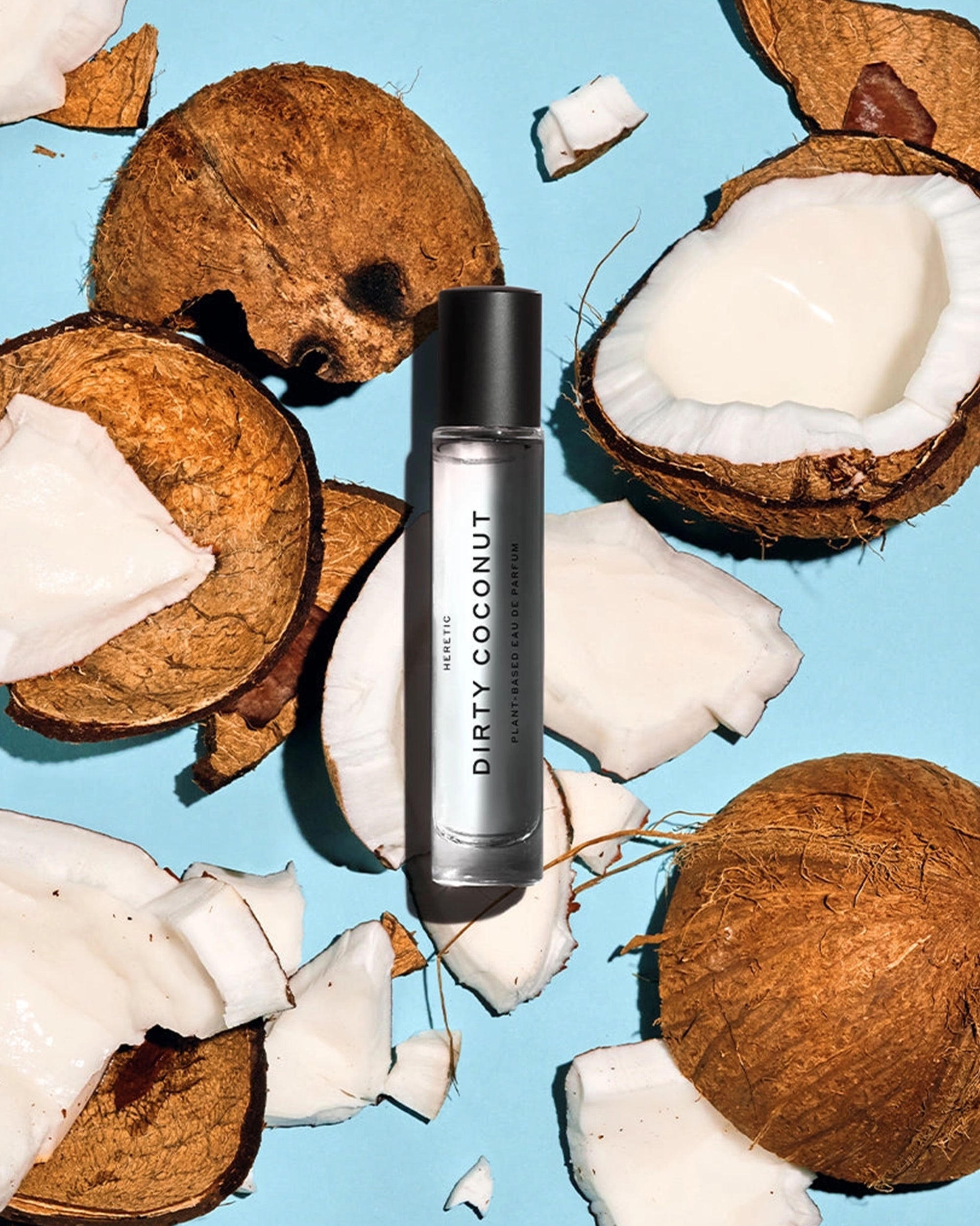 A bottle of Faire Heretic 15ML Perfume rests on a blue surface, surrounded by cracked open coconuts and pieces of coconut flesh.