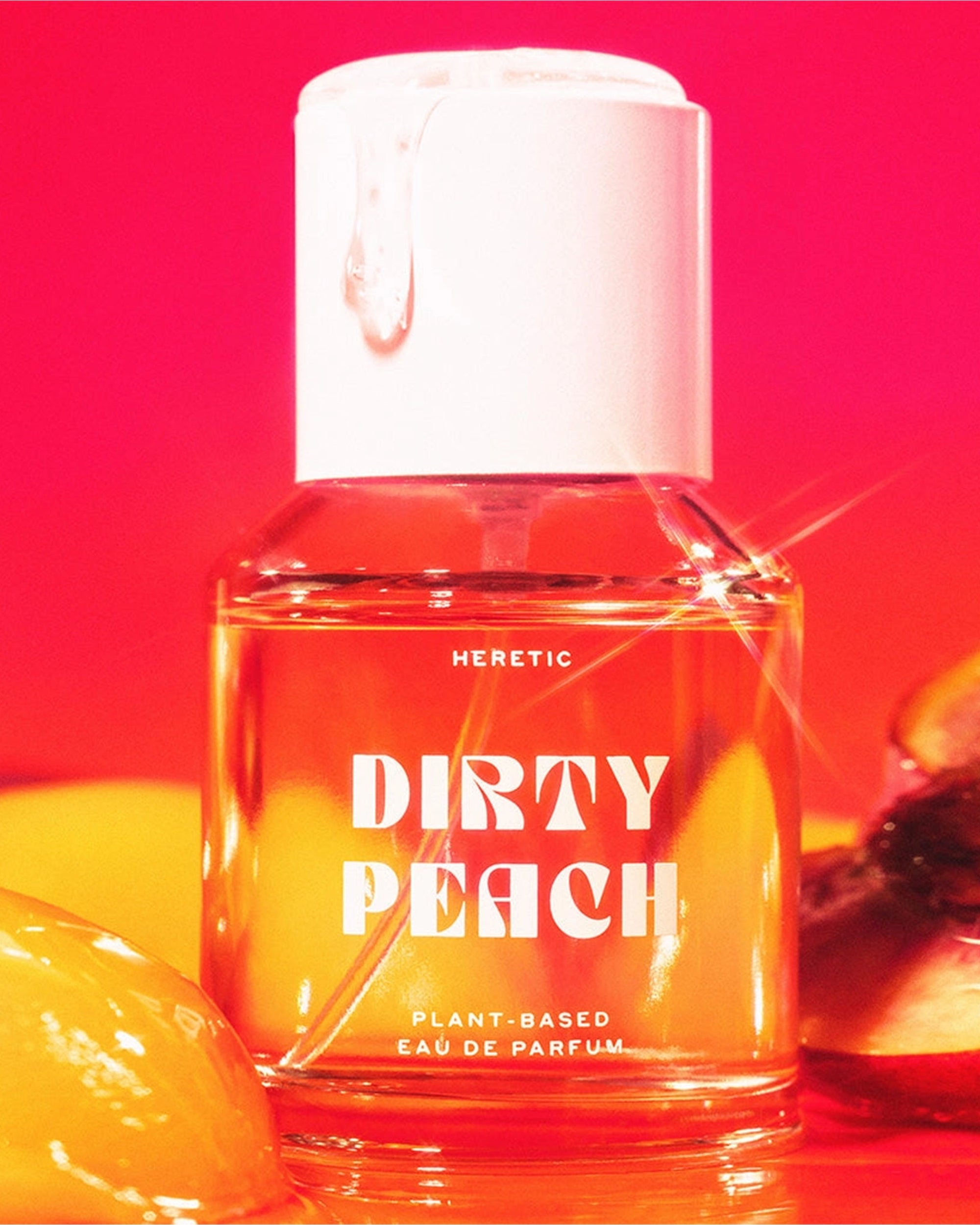 A clear bottle of Heretic 50ML Perfume by Faire, labeled "Dirty Peach," rests against a vibrant red and orange background with peach slices and syrup, suggesting a sensual fragrance featuring sandalwood vanilla notes.