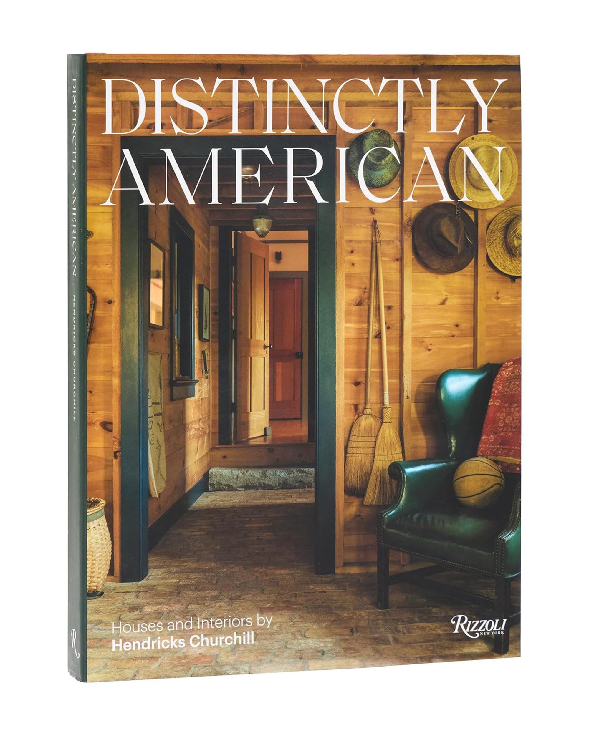 The book cover of "Distinctly American" by Random House showcases vernacular architecture—a rustic wooden hallway with hats and tools on the wall, a green leather armchair, and a view into another room.