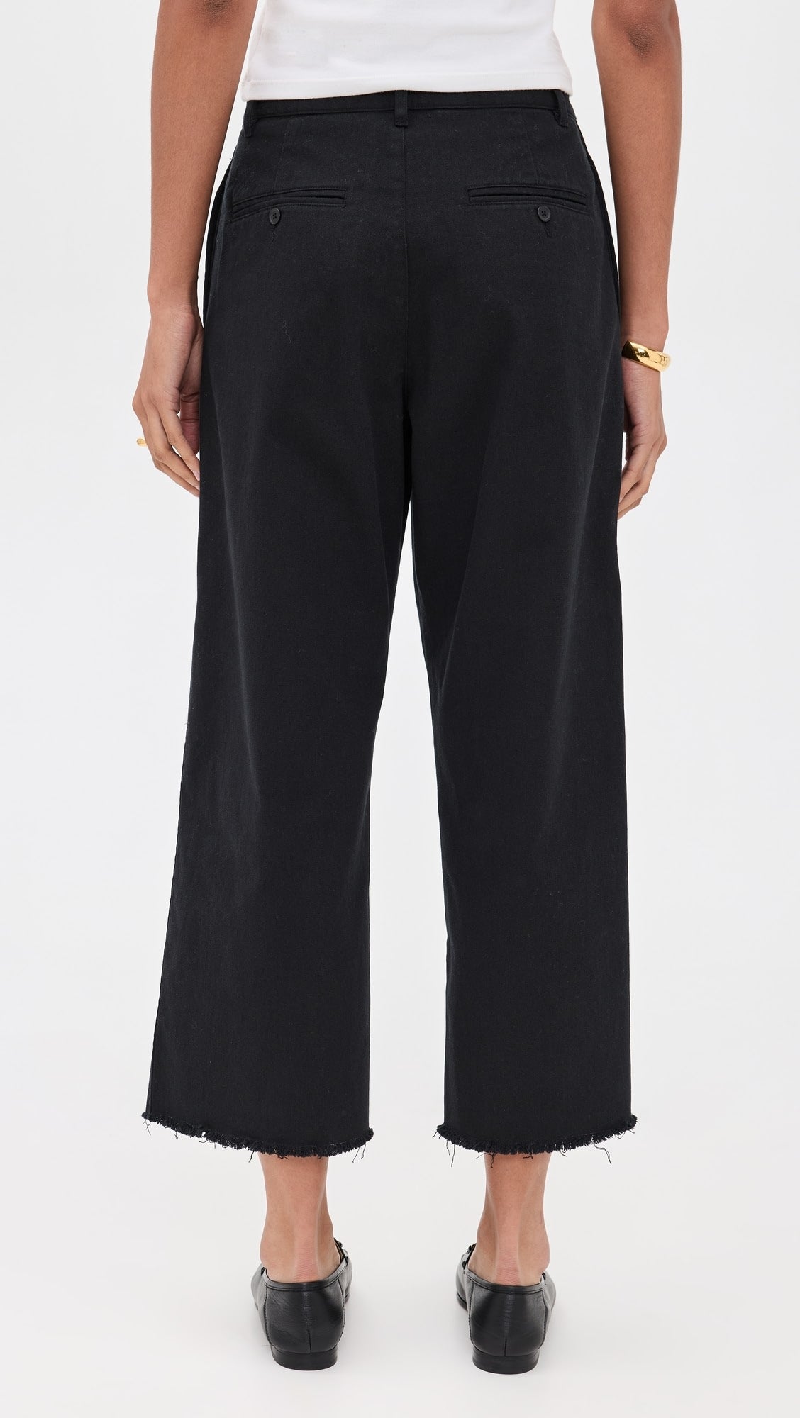 A person wears Denimist's CROPPED DOUBLE PLEAT WIDE LEG CHINO in black 100% cotton with raw hems, paired with a white top, black shoes, and gold jewelry, shown from the back against a plain white background.