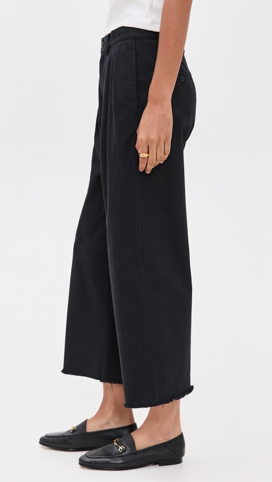 A person wearing Denimist CROPPED DOUBLE PLEAT WIDE LEG CHINO in black, paired with a white top, black loafers, and gold rings, stands against a plain light background.