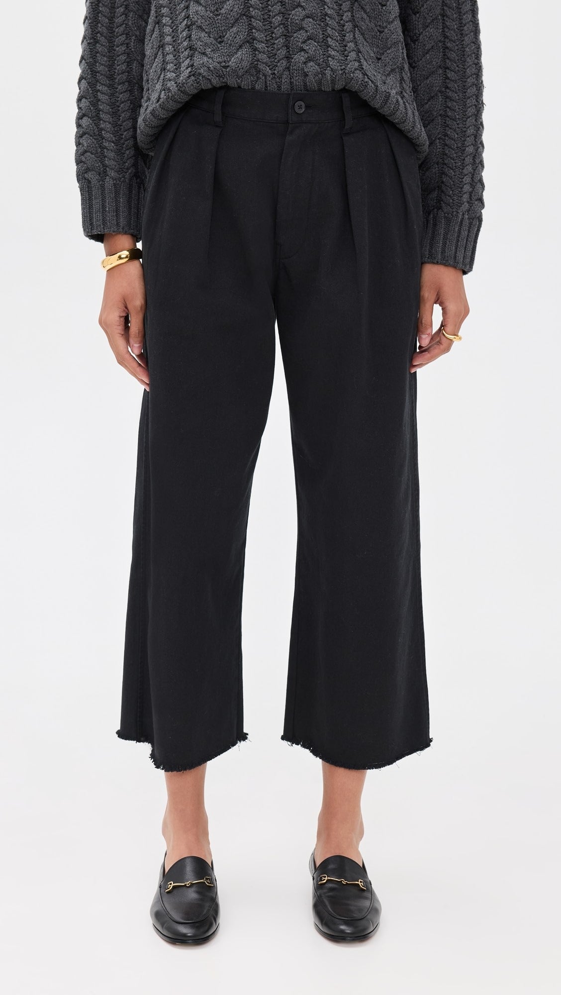 A person stands against a plain white background wearing a chunky gray knit sweater, Denimist CROPPED DOUBLE PLEAT WIDE LEG CHINO pants in black, black loafers, and gold bracelets.