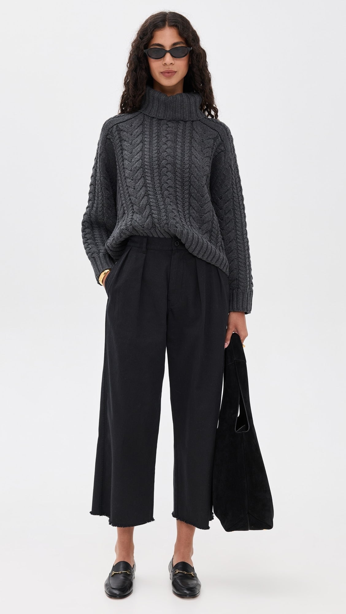 A woman stands against a plain white background wearing a chunky dark gray cable knit turtleneck, Denimist CROPPED DOUBLE PLEAT WIDE LEG CHINO pants in black, black loafers, sunglasses, and holding a black tote bag.