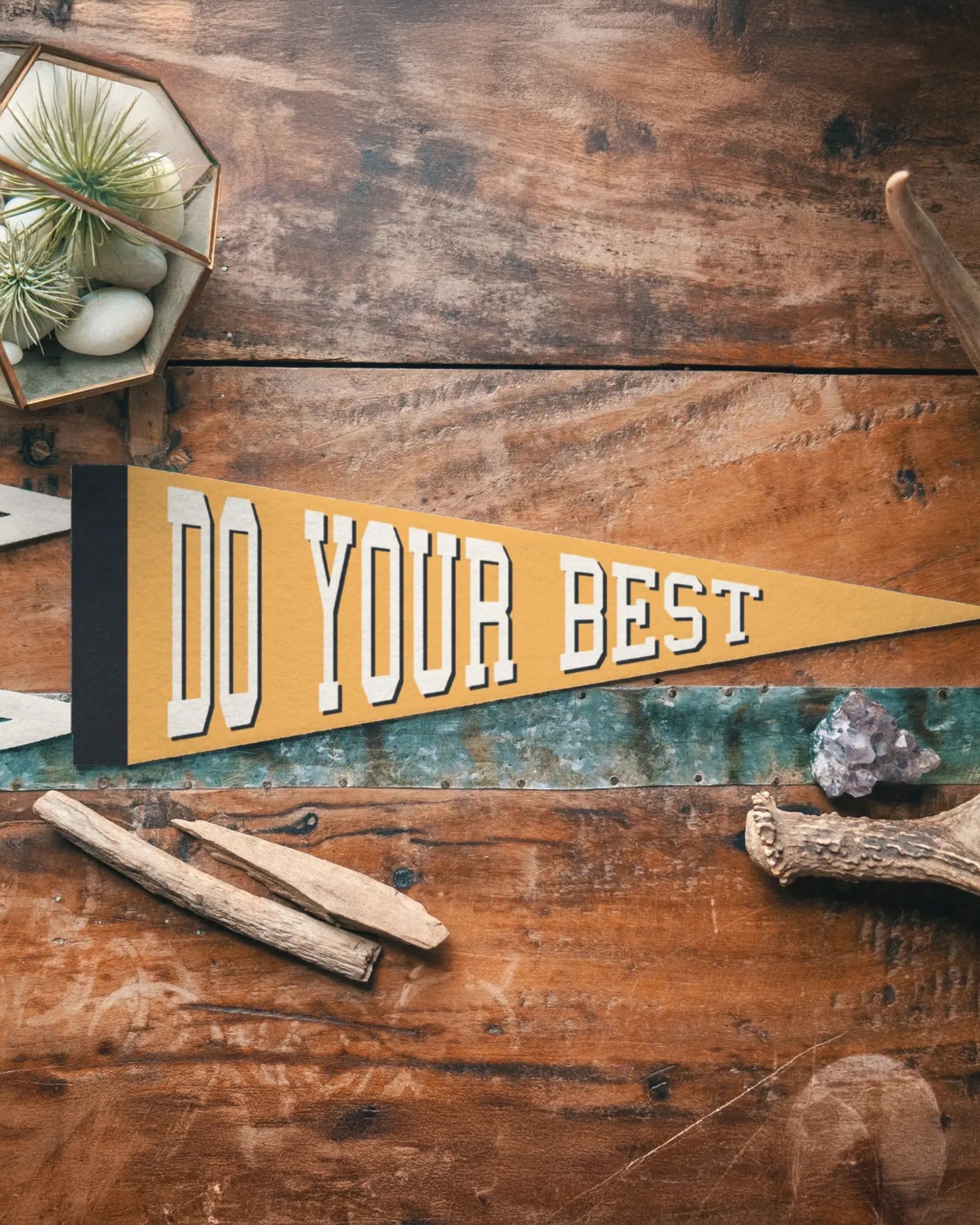 The "Do Your Best" Felt Pennant by Faire features bold white text on yellow felt and is displayed on a rustic wooden table among plants and rocks—ideal for vintage sports pennant fans or as kids room decor.