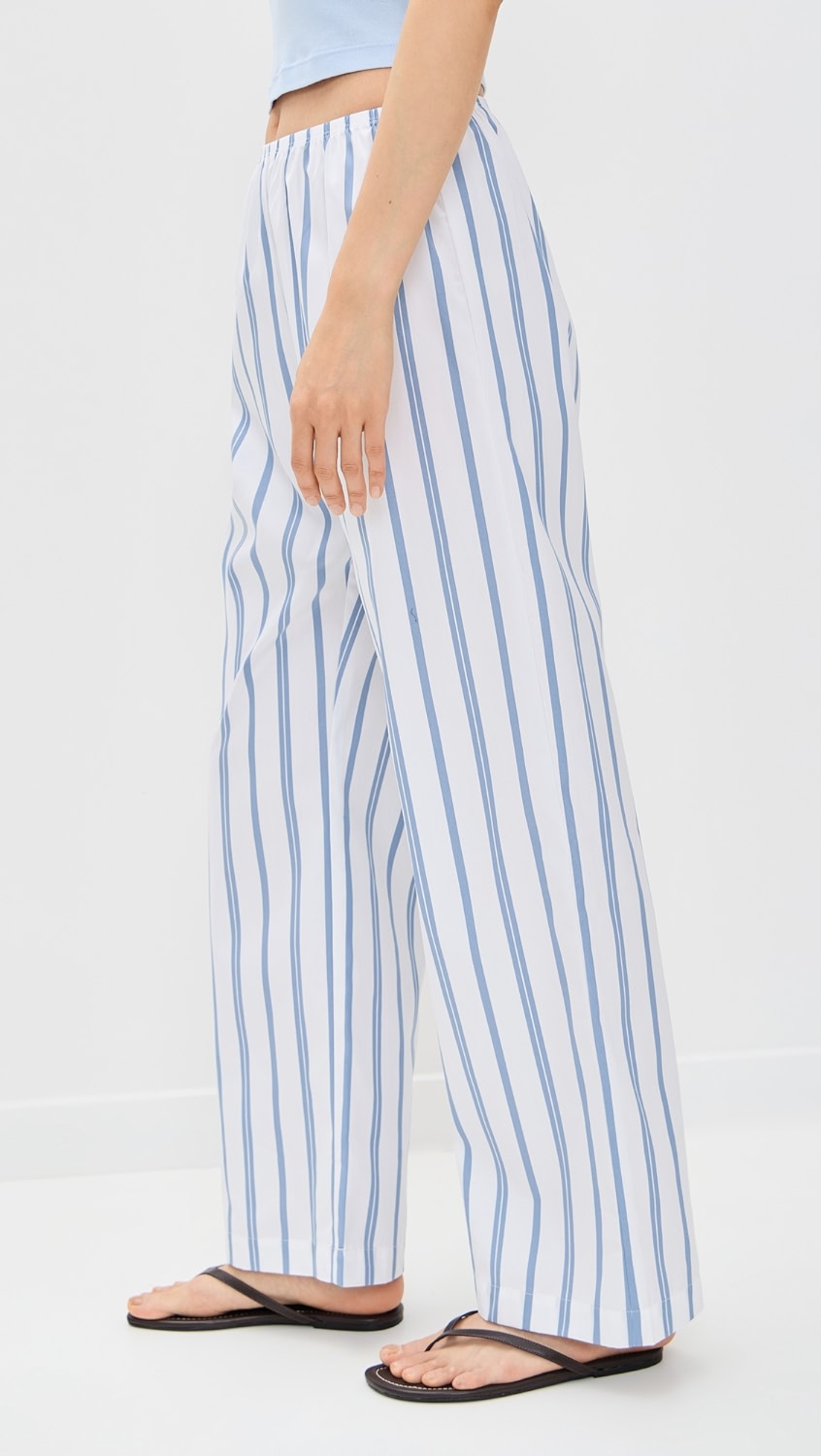 Wearing Donni’s The Pop Simple Pant in white stripe—a loose, lightweight poplin style—paired with a light blue top and black flip-flops, the lower half of the body is shown against a plain white background.