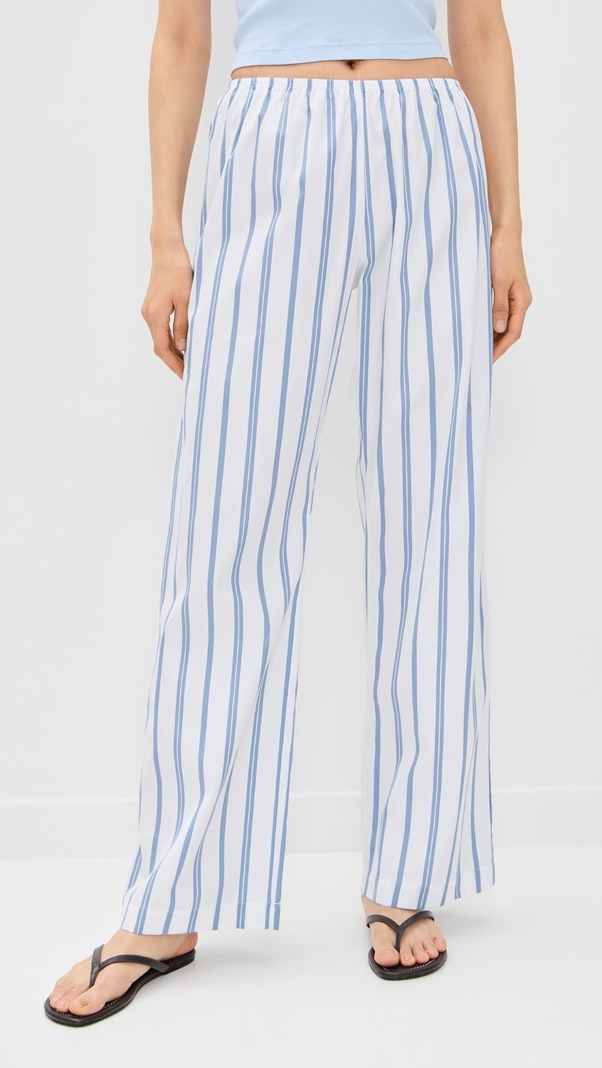 A person models Donni's The Pop Simple Pant—loose-fitting white cotton pants with vertical blue stripes—paired with a light blue top and black flip-flops, standing against a plain white background.
