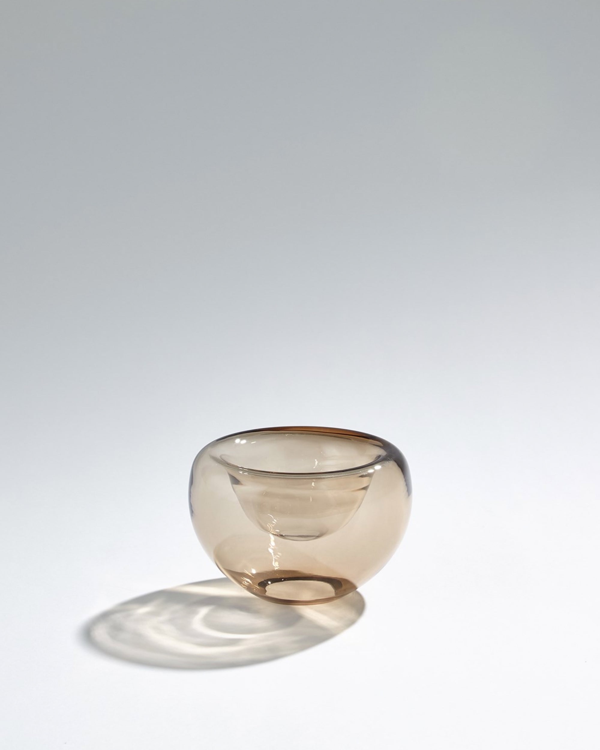 The ASH6.60035 Double Take Bowl SM by Global Views rests on a white surface, its small, round shape in translucent topaz glass casting soft shadows and subtle reflections against a minimalist background.