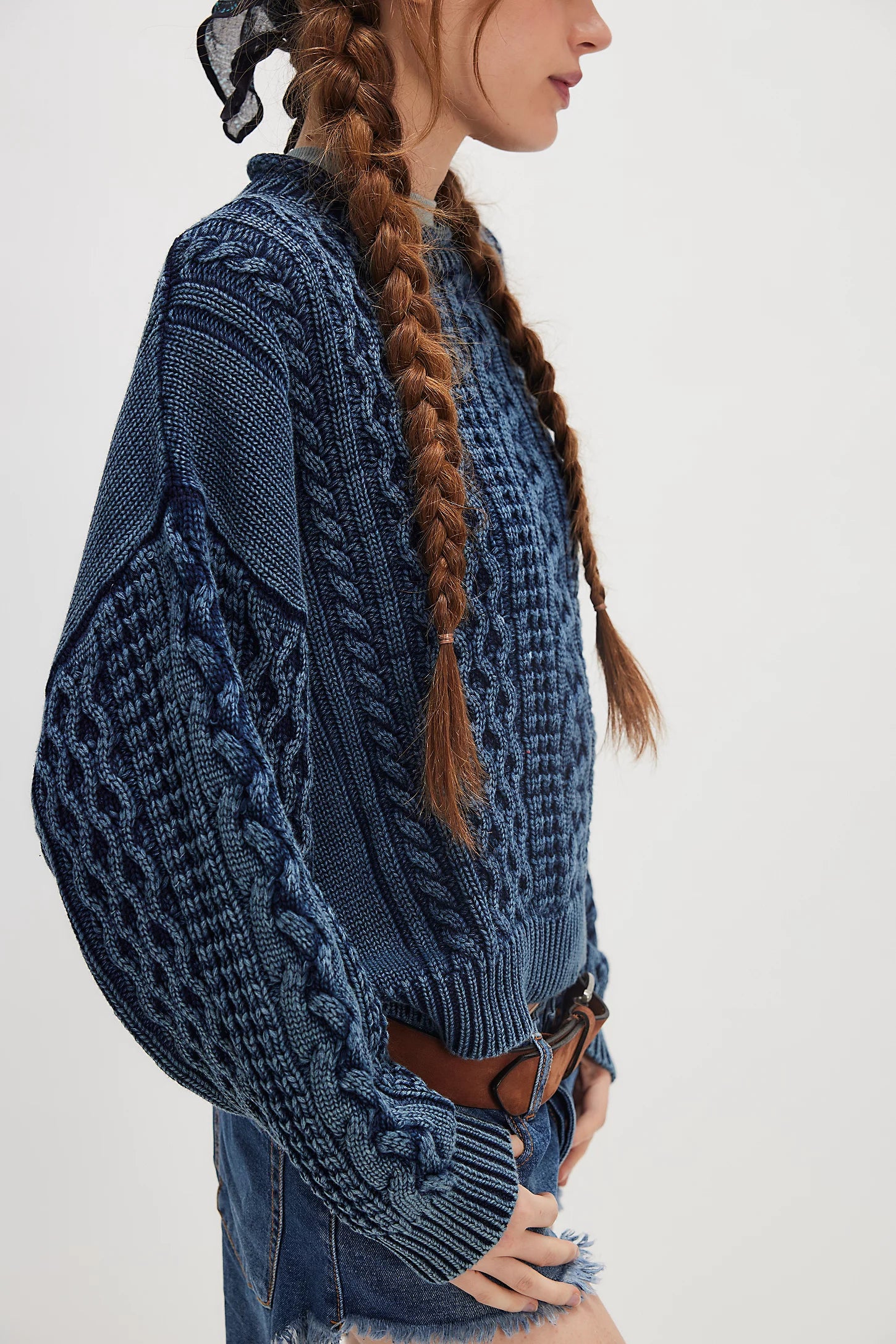 A person with long braided hair wears the Denimist OVERSIZED ARAN SWEATER W/ROLLED NECK and denim shorts with a brown belt, viewed from the side against a plain light background for a cozy, timeless look.