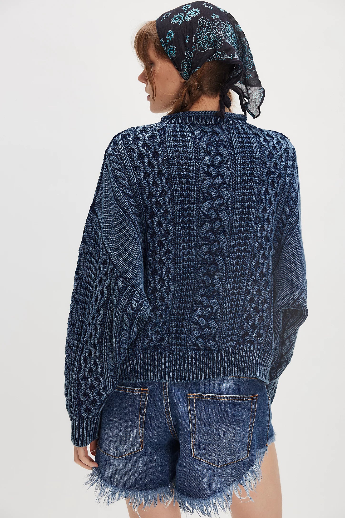A person shown from the back wears the Denimist OVERSIZED ARAN SWEATER W/ROLLED NECK in dark blue, paired with frayed denim shorts and a black scarf with blue patterns, set against a plain light background for a relaxed heritage vibe.