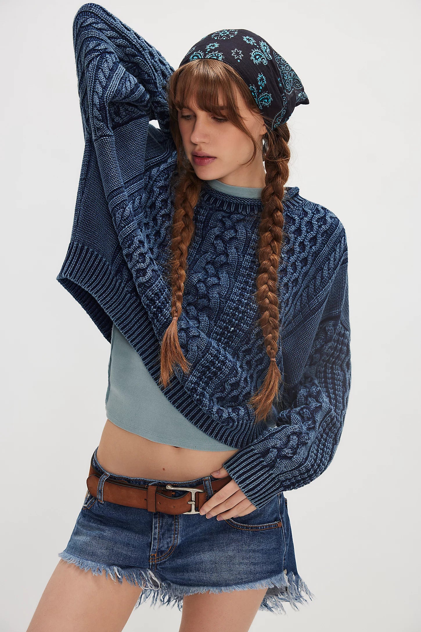 A young woman with long brown braids and a patterned bandana wears the Denimist OVERSIZED ARAN SWEATER W/ROLLED NECK over a light blue top, styled with denim shorts and a brown belt, posing with one arm bent and eyes closed.