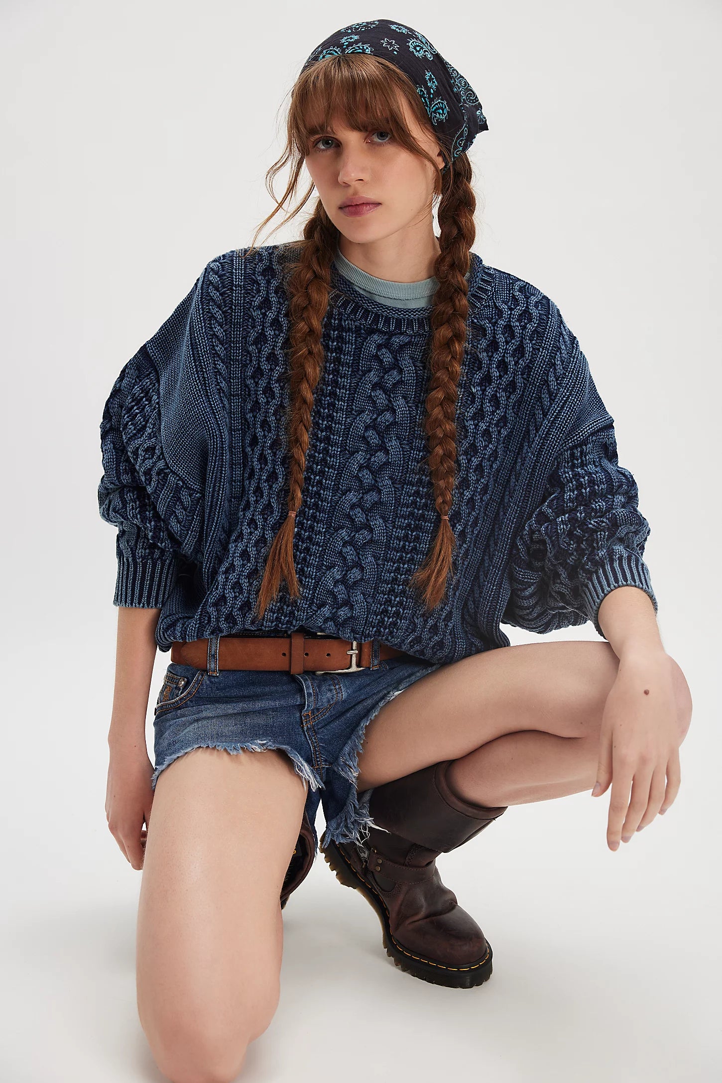 A young woman wearing the Denimist OVERSIZED ARAN SWEATER W/ROLLED NECK, styled with denim shorts, a brown belt, dark boots, and a patterned headscarf kneels against a plain light background for a cozy heritage look.