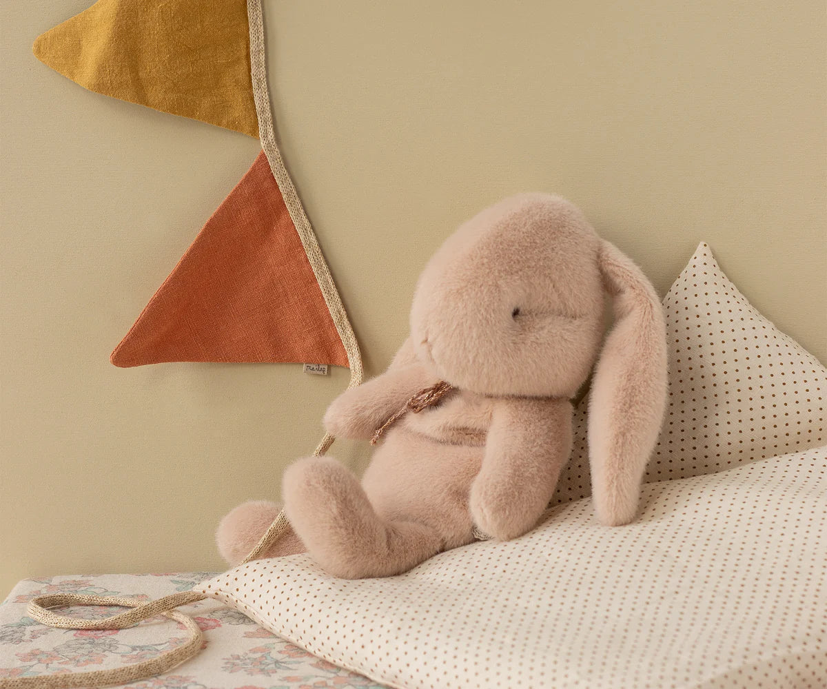 The Maileg Bunny Plush, with floppy ears, rests on a polka-dot pillow beside a wall adorned with mustard yellow and rust fabric pennants, creating a cozy and gentle atmosphere.