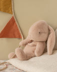 The Maileg Bunny Plush, with floppy ears, rests on a polka-dot pillow beside a wall adorned with mustard yellow and rust fabric pennants, creating a cozy and gentle atmosphere.