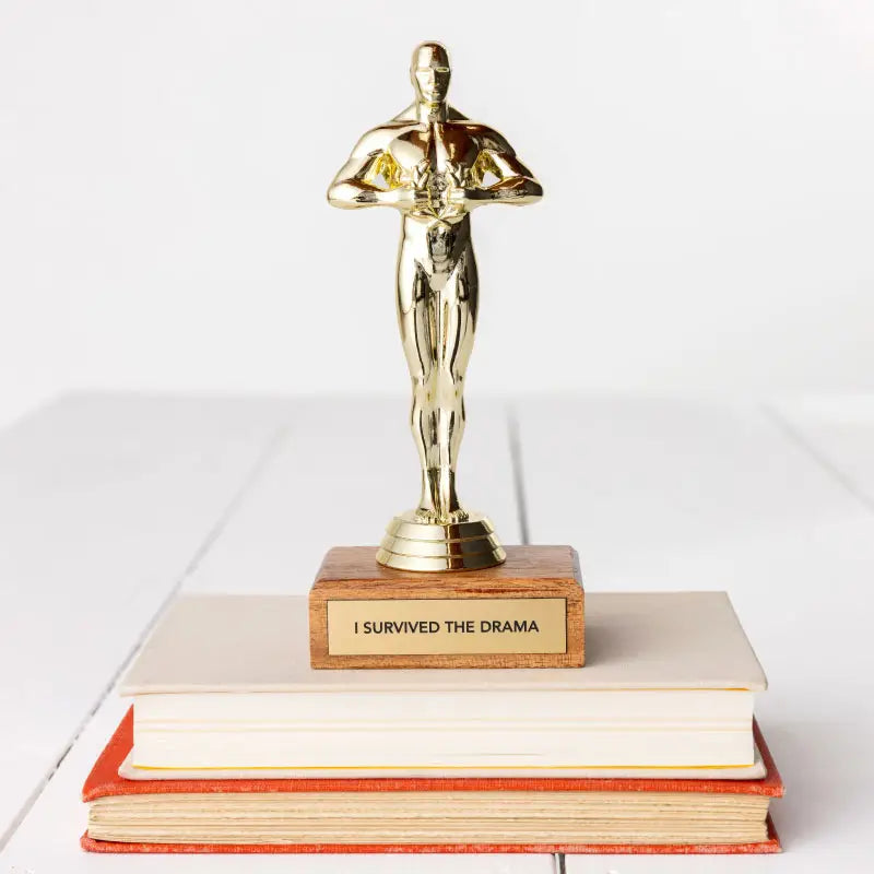 A JE Trophy resembling an Oscar statuette stands on top of stacked books, with a plaque reading &quot;I SURVIVED THE DRAMA.&quot; The background is a plain white surface in Arizona-style.