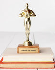 A JE Trophy resembling an Oscar statuette stands on top of stacked books, with a plaque reading "I SURVIVED THE DRAMA." The background is a plain white surface in Arizona-style.
