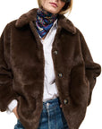 Model wears the Alex Mill Duane St Jacket in dark brown faux fur with front snaps over a white shirt and blue jeans, accessorized with a colorful scarf and sunglasses, standing against a plain white background.