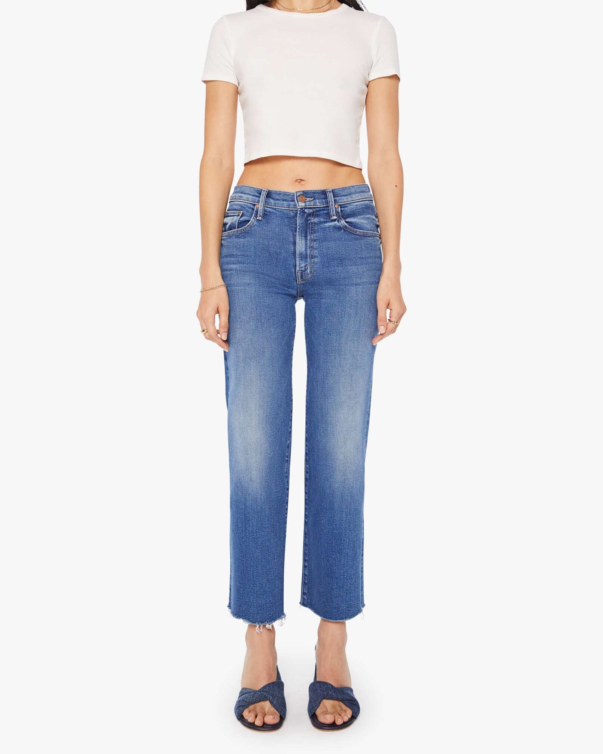 A woman stands facing forward against a white background, wearing Mother’s The Mid Rise Rambler Zip Ankle Fray jeans, a short-sleeved white crop top, and dark blue slip-on sandals.