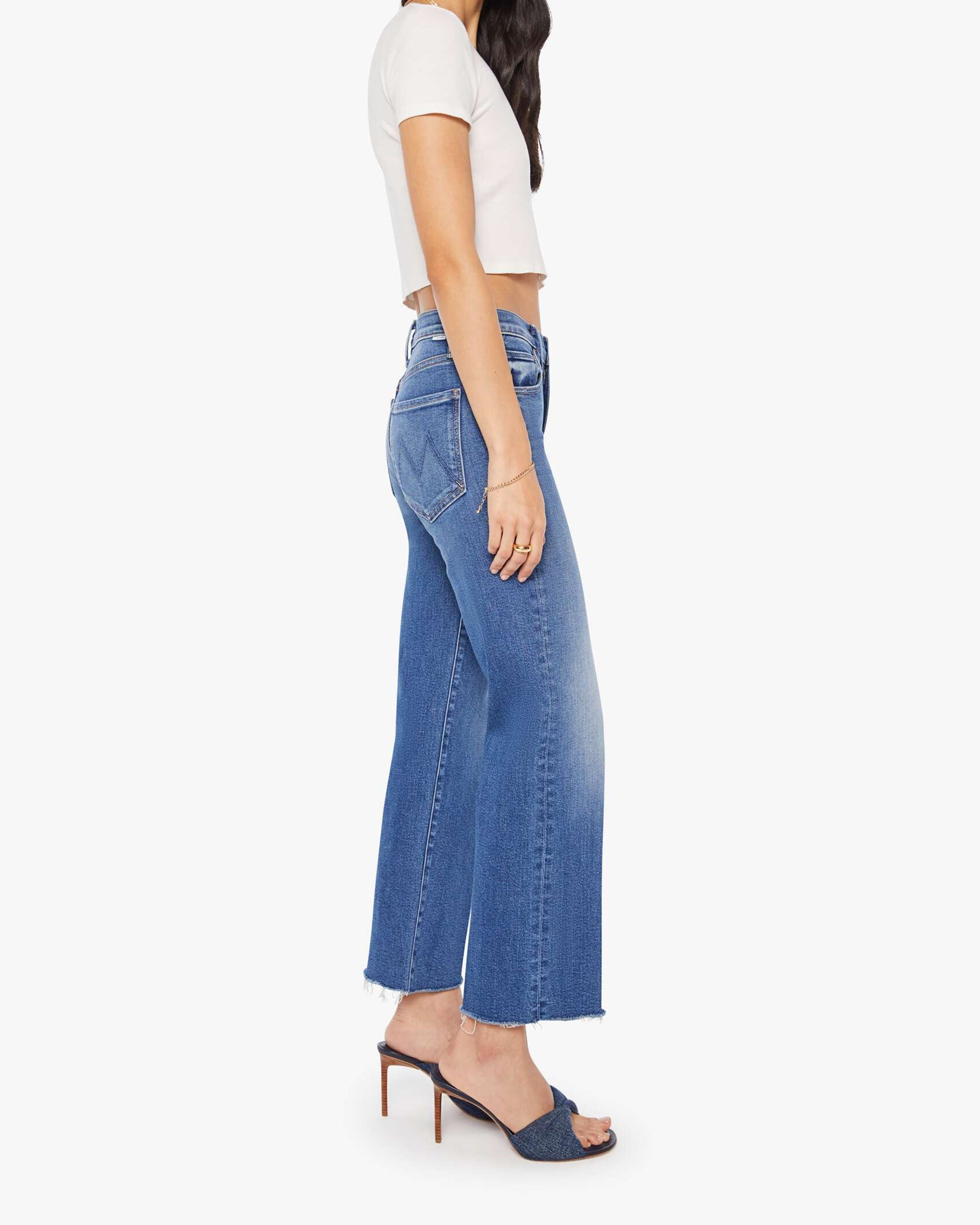 A woman stands sideways against a white background wearing Mother’s The Mid Rise Rambler Zip Ankle Fray jeans, a white short-sleeve crop top, and dark high-heeled sandals.