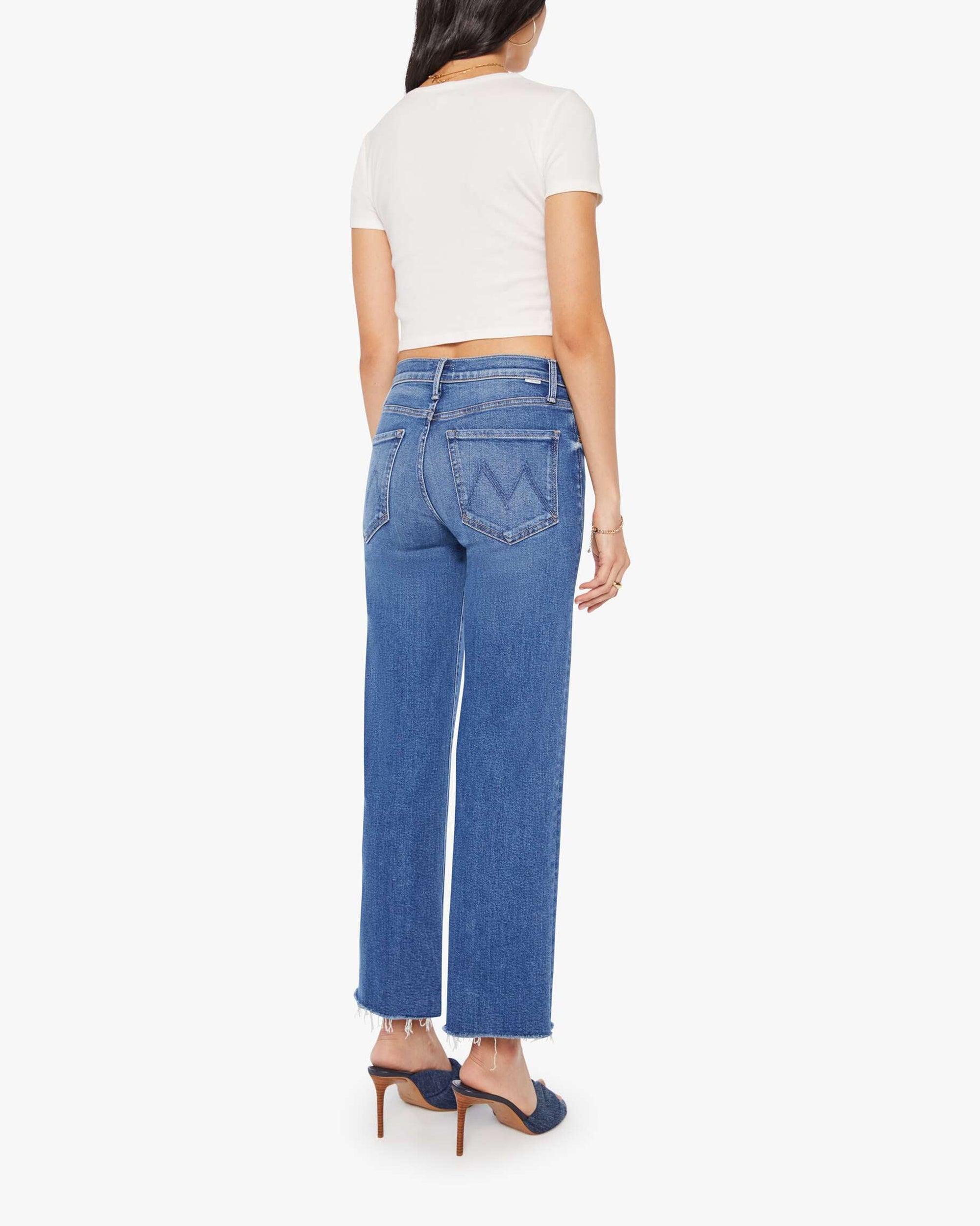 A woman wears Mother’s The Mid Rise Rambler Zip Ankle Fray jeans, a white short-sleeve top, and blue heeled shoes, standing with her back to the camera against a plain white background.