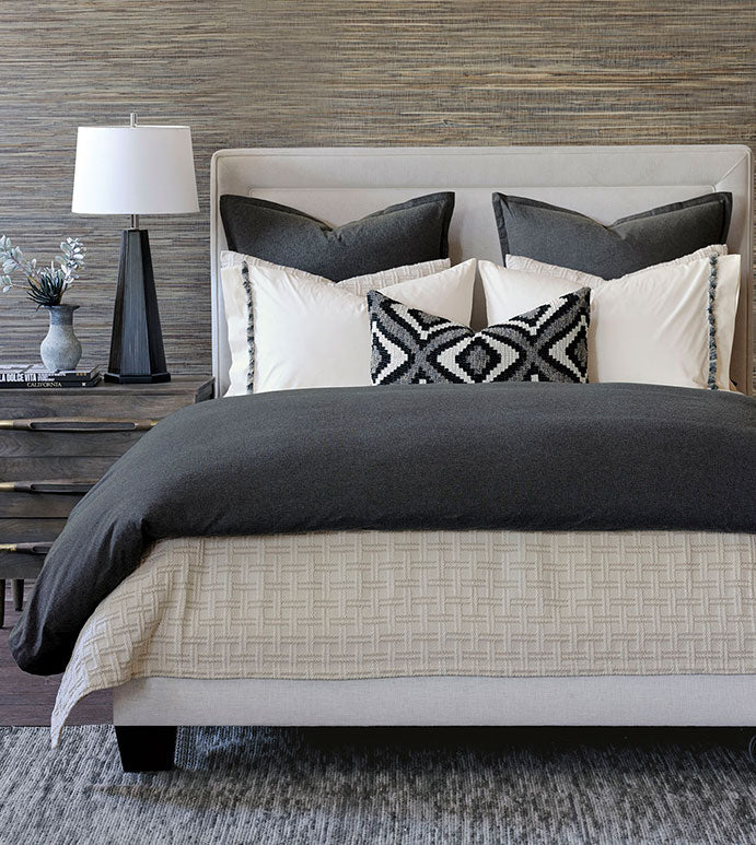 A neatly made bed showcases the Eastern Accents Monterosa Cotton Flannel Duvet Cover in dark gray with a hidden zipper, black, white, and gray pillows, a cream textured bedspread, and a bedside table with lamp and vase on the left.