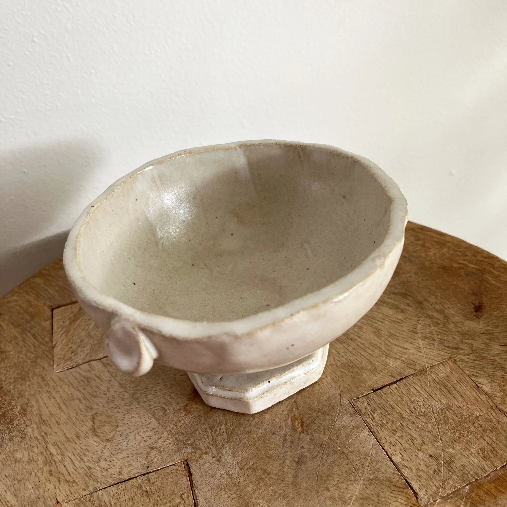 The Tiny Trophy Bowl by Faire is a handmade, off-white glazed stoneware piece featuring an uneven rim and small circular handle, displayed on wood against a white wall—a unique sugar dish.