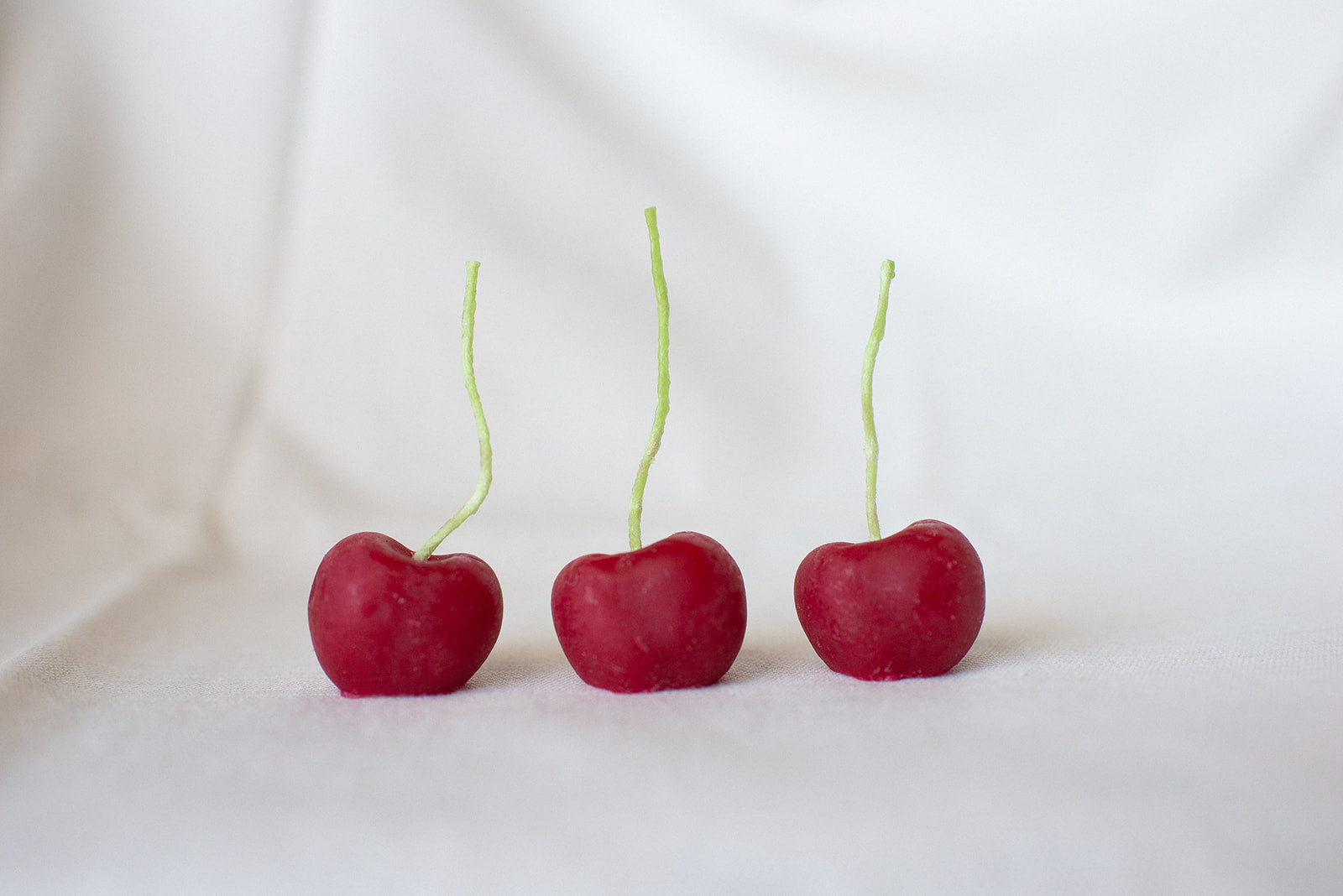 Faire’s Cherry Beeswax Candles feature three upright, cherry-shaped candles with green stems and a vivid red color, beautifully resembling real cherries on a white surface.