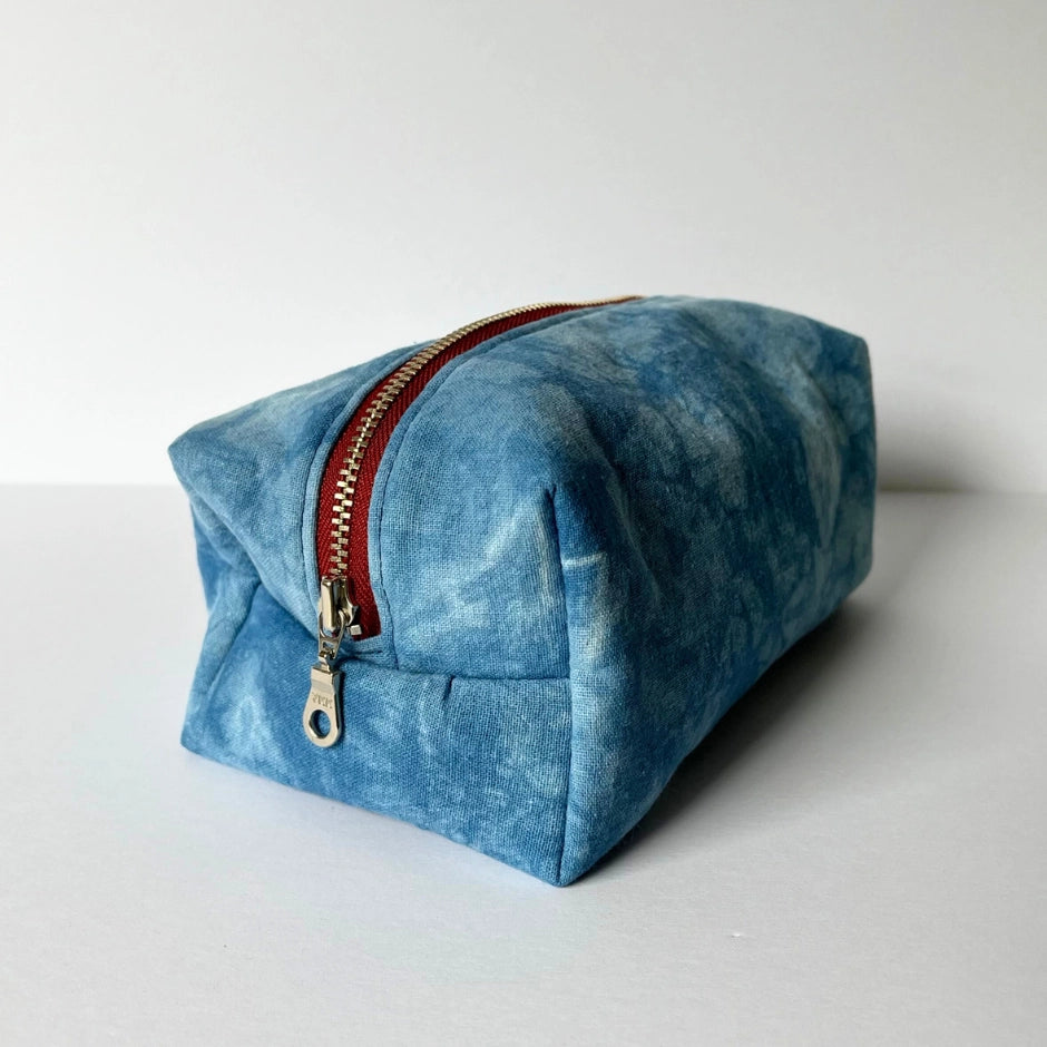 The Faire Dyed Toiletries Bag is a rectangular blue cotton pouch with a hand-dyed marbled design and a red zipper with metal pull tab, shown on a white surface—ideal for organizing your desk.