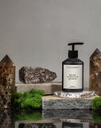 A bottle of Faire Inspired Hand Soap rests on a stone slab amid moss, greenery, two brown crystal towers, and rocks, featuring a diptyque-inspired touch against a neutral gray background.