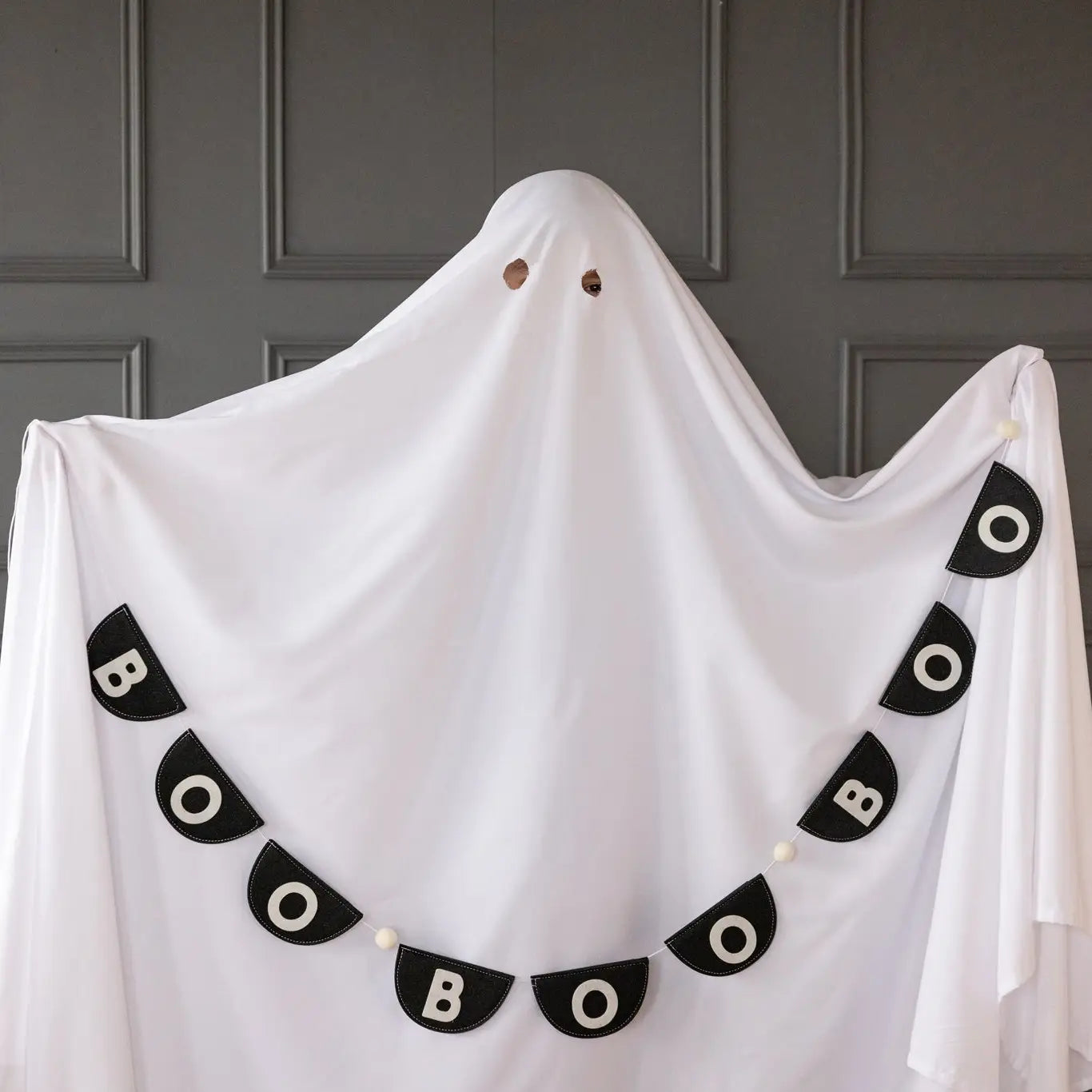 A person dressed as a ghost stands against a gray wall, with the My Mind's Eye Bunting Banner hanging in front. The banner features black fabric and white letters spelling "BOO BOO BOO.
