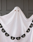A person dressed as a ghost stands against a gray wall, with the My Mind's Eye Bunting Banner hanging in front. The banner features black fabric and white letters spelling "BOO BOO BOO.