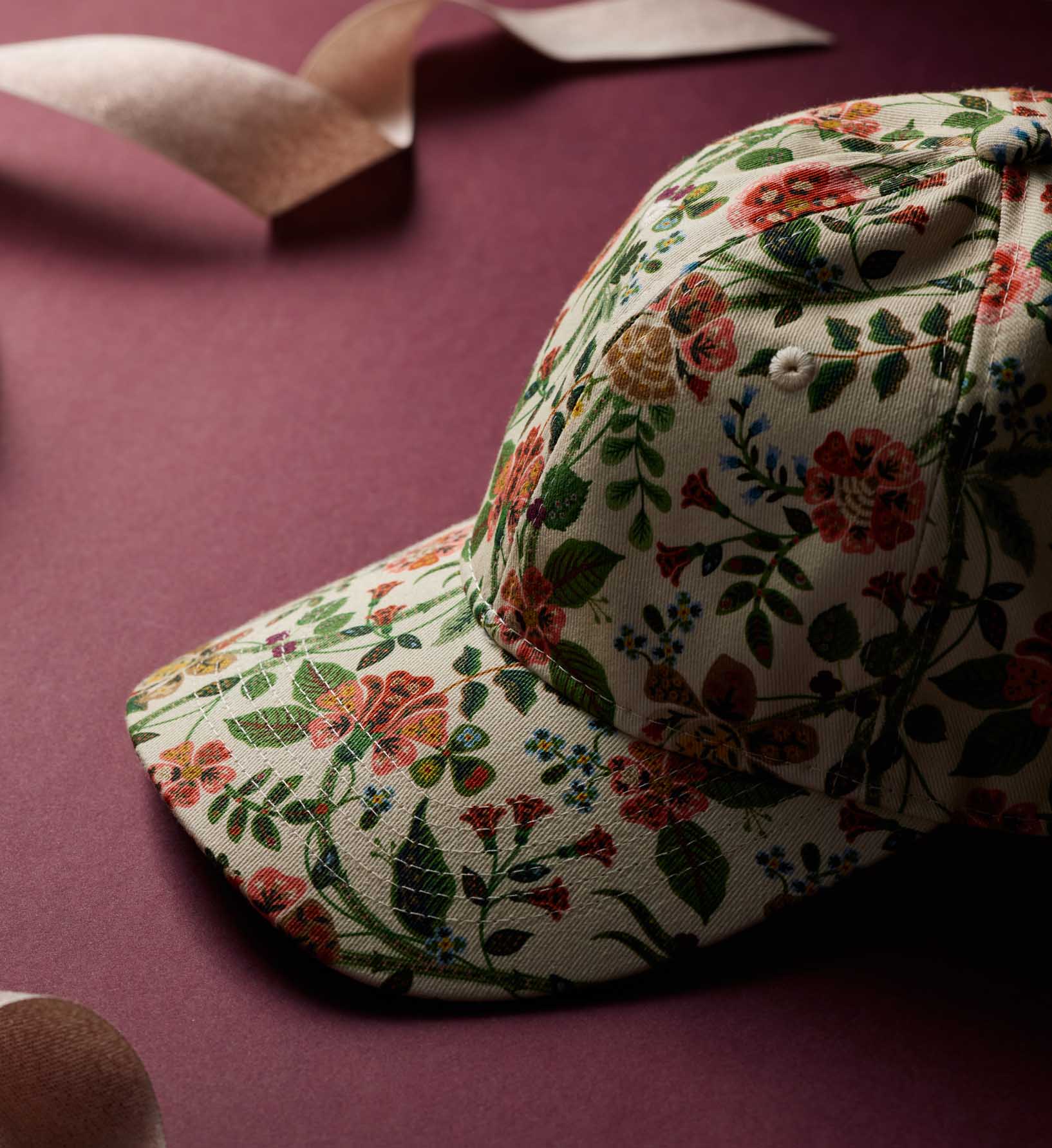 The Gracie Baseball Cap by Faire features a vibrant white floral print with red, pink, and green accents. Shown on a maroon surface with matching ribbon accents in the background, it offers an adjustable strap for a comfortable fit.