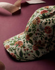 The Gracie Baseball Cap by Faire features a vibrant white floral print with red, pink, and green accents. Shown on a maroon surface with matching ribbon accents in the background, it offers an adjustable strap for a comfortable fit.
