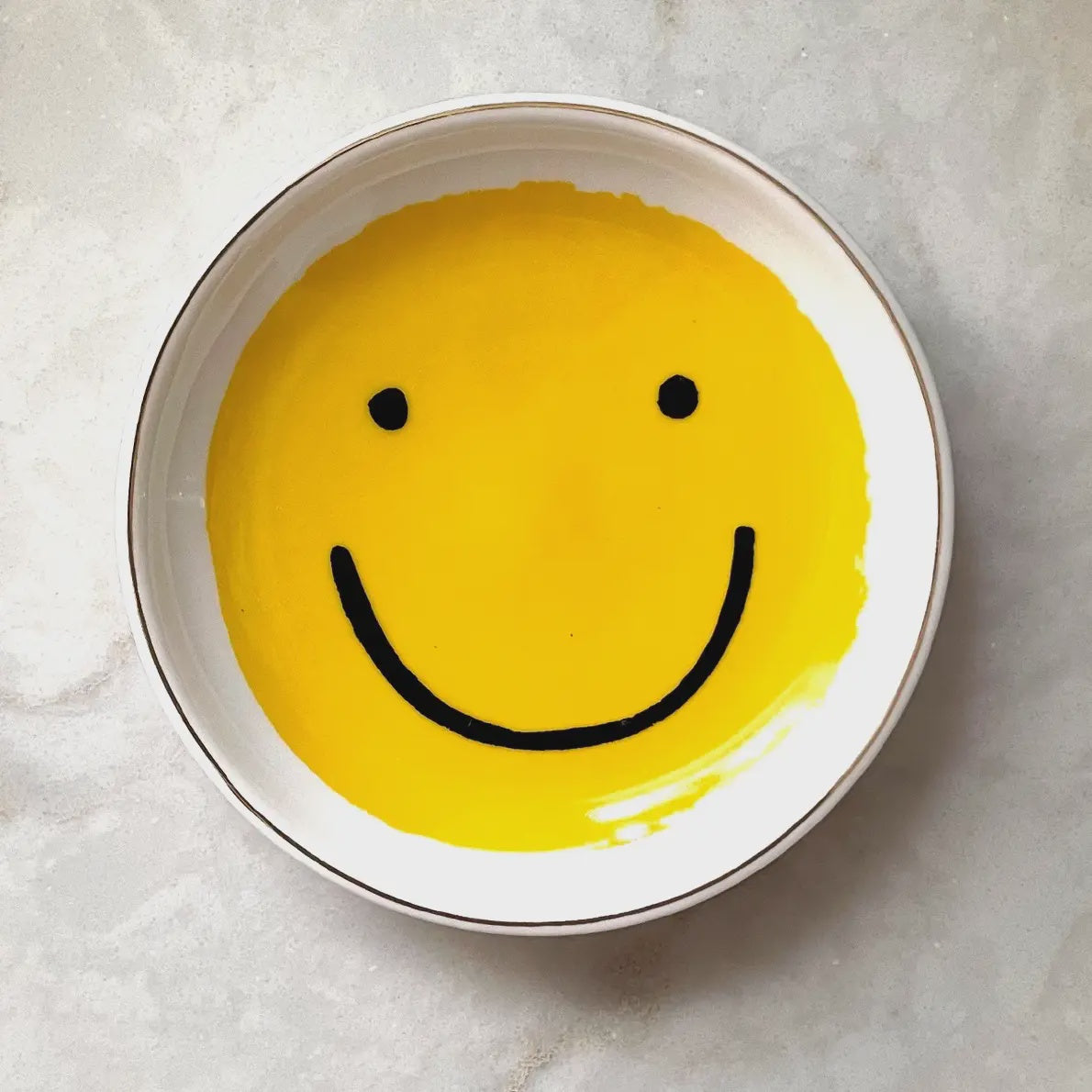 A round, ceramic Trinket Dish by Faire showcases a prominent yellow smiley face with black eyes and a wide smile. It is set on a light gray, textured surface.