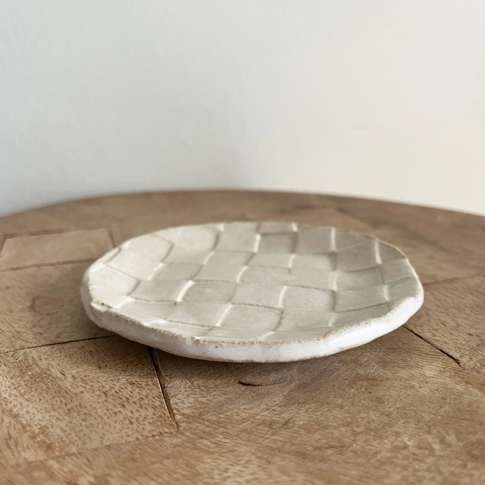 The Faire Ghost Checker Trinket Dish, a small, round glazed stoneware dish with a woven checkerboard pattern, rests on a wooden surface against a plain, light-colored background.