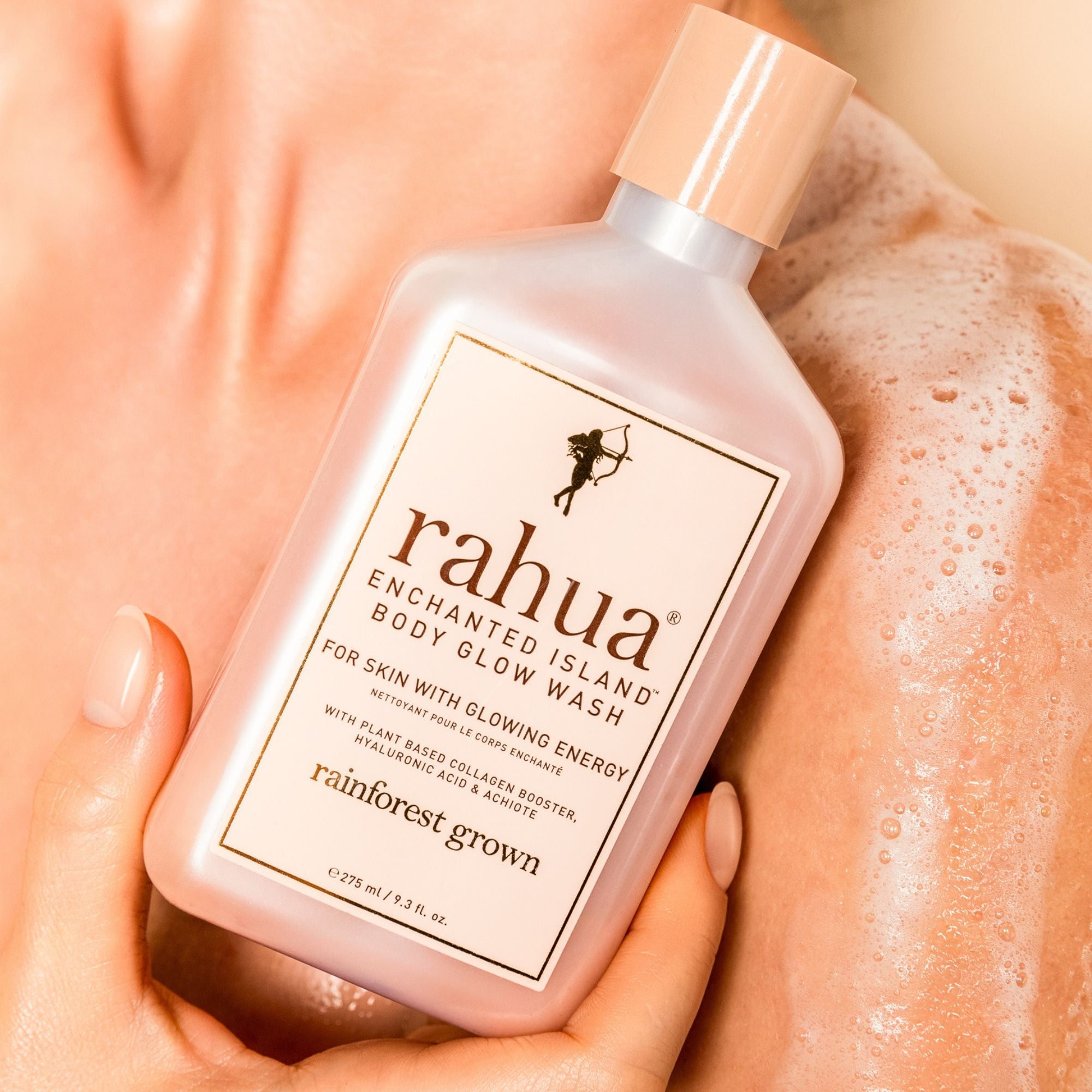 A close-up shows a person holding a pink bottle of Faire Rahua Enchanted Island Body Glow Wash against their soapy shoulder, with the moisturizing body wash’s label clearly visible.