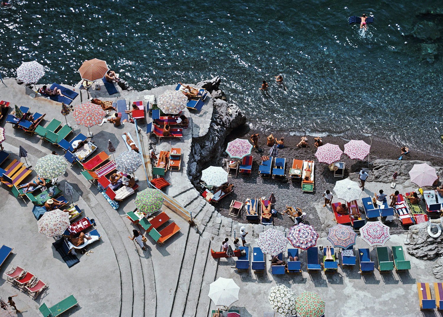 Enjoy a vibrant, midcentury beach scene with the Pieceworks Puzzle 1000 by Faire—featuring colorful umbrellas, lounge chairs, sunbathers on a terrace, and swimmers in crystal-clear blue water.