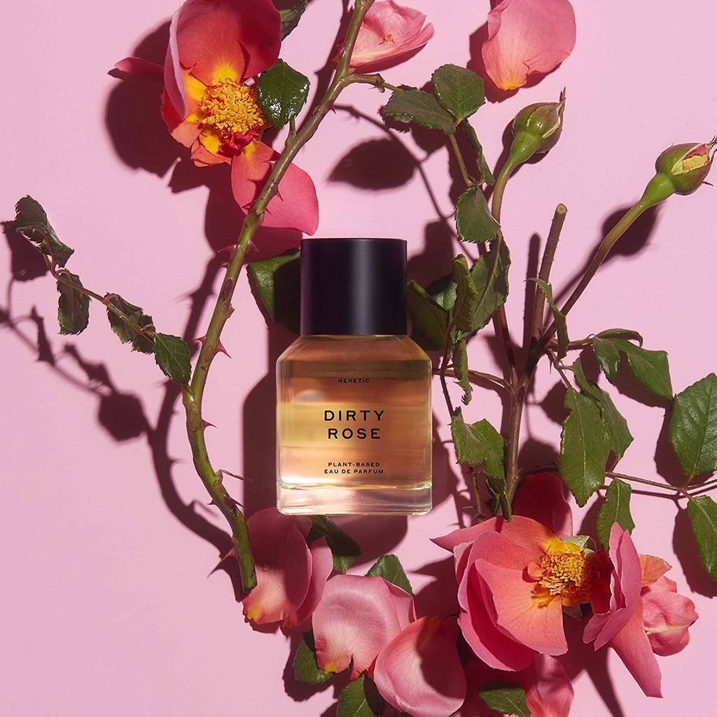 A bottle of Faire Heretic 50ML Perfume, labeled “Dirty Rose,” is displayed amid pink rose flowers and green leaves on a pink background, evoking a sensual fragrance.