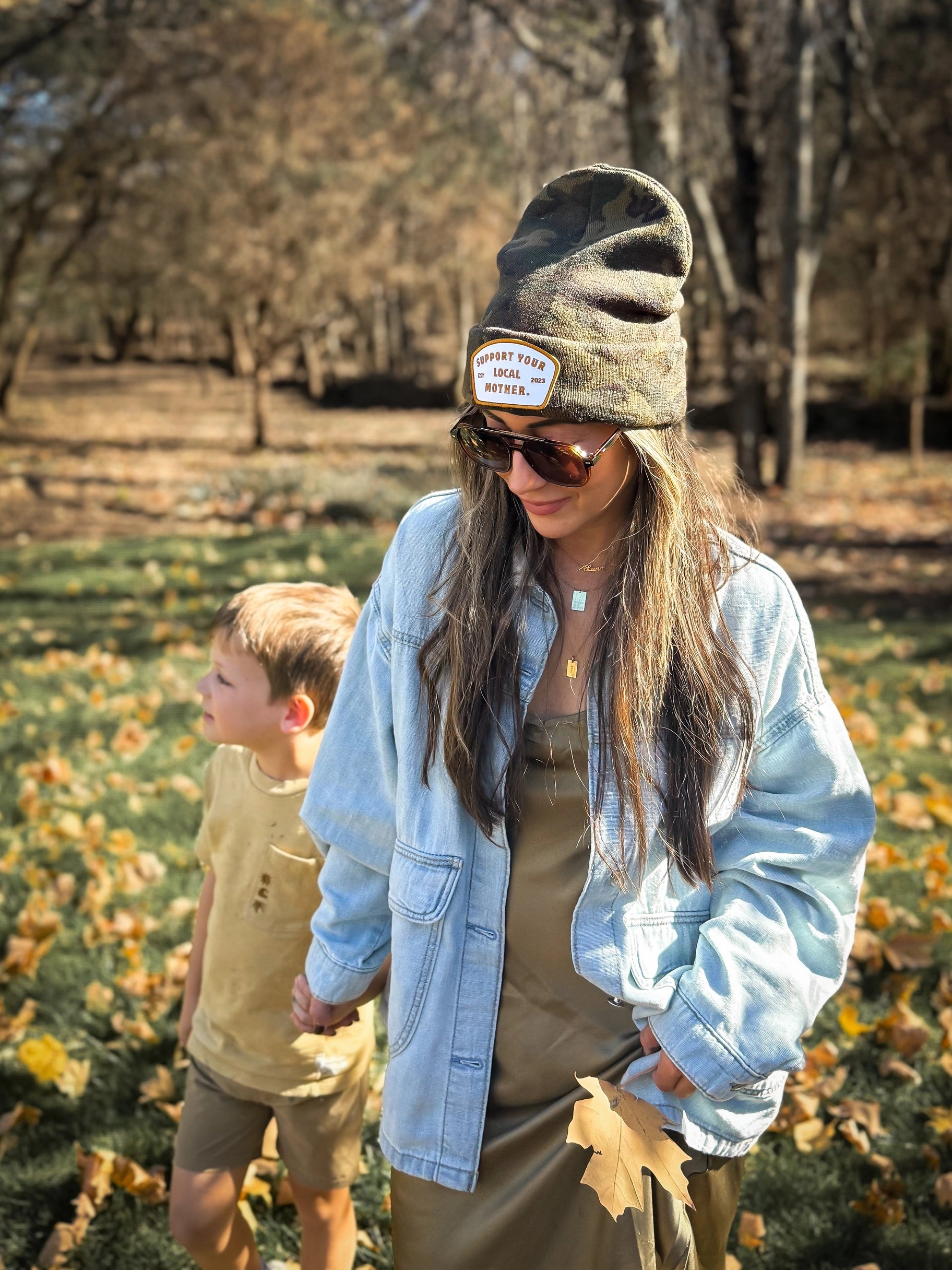 A woman in Faire’s Patch Beanie Support Your Local Mother, sunglasses, and a light denim jacket holds hands with a young boy in tan as they walk outdoors on leaf-covered grass with trees in the background.